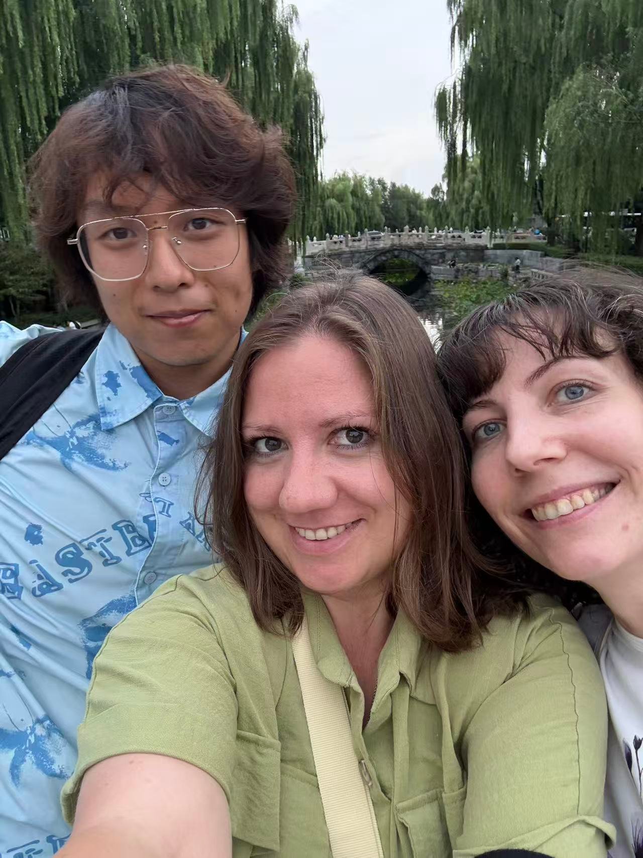 我们的合照 画面中有三个人正在合影,左侧是一名戴眼镜的长发男子,身穿浅蓝色印花衬衫。中间是一名棕色中长发的女子,身穿绿色上衣,面带微笑。右侧是一名深色短发的女子,露出牙齿微笑,背景是垂柳和一座石拱桥。