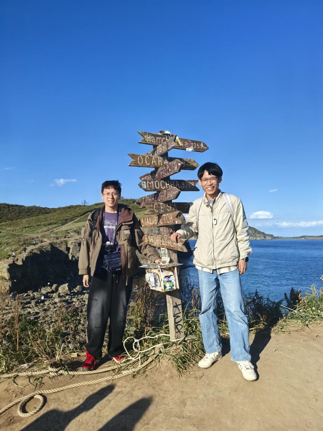 Two people standing by the sea under a blue sky, with a signpost showing distances to various cities behind them. The person on the left wears a brown coat and black pants, while the person on the right wears a light-colored jacket and blue jeans.