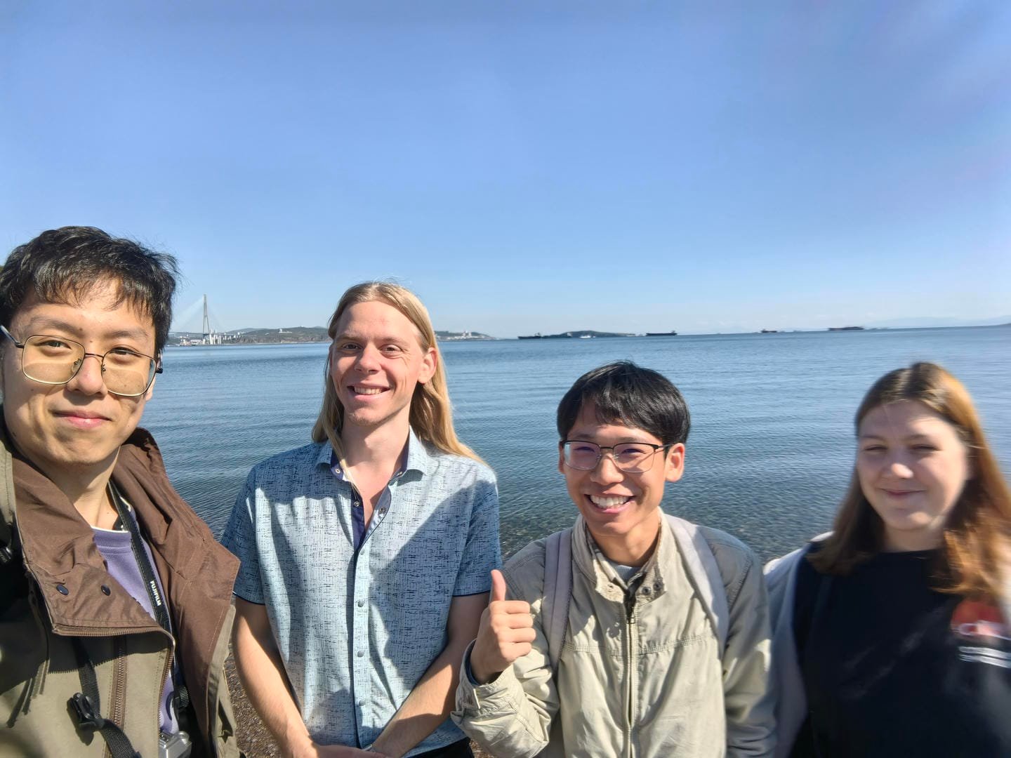 Four people standing by the sea under a blue sky. From left to right: the first person wears glasses and a brown coat; the second has long blonde hair and a blue shirt; the third wears glasses, a light-colored jacket, and gives a thumbs-up; the fourth has brown hair and a dark top.