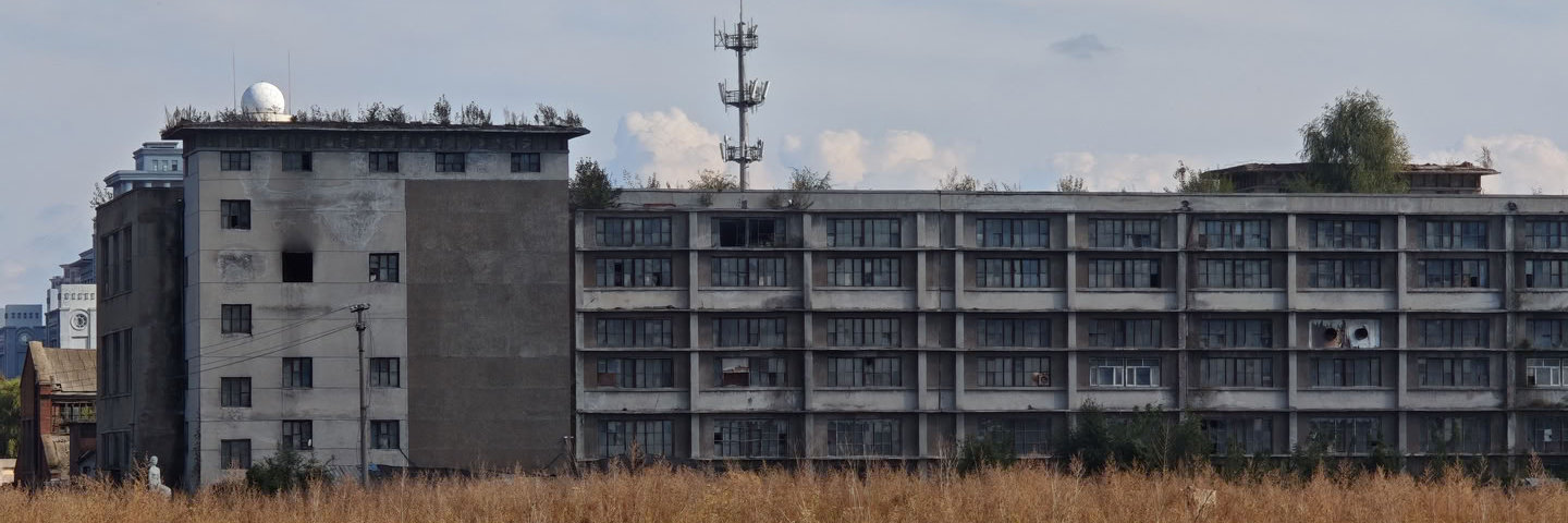 一座长条形的灰色多层建筑横跨画面，窗户排列整齐但部分破损。建筑左侧连接着一栋较高的楼房，屋顶设有白色球状物体。建筑后方矗立着一座通信铁塔，前景是大片枯黄的杂草。