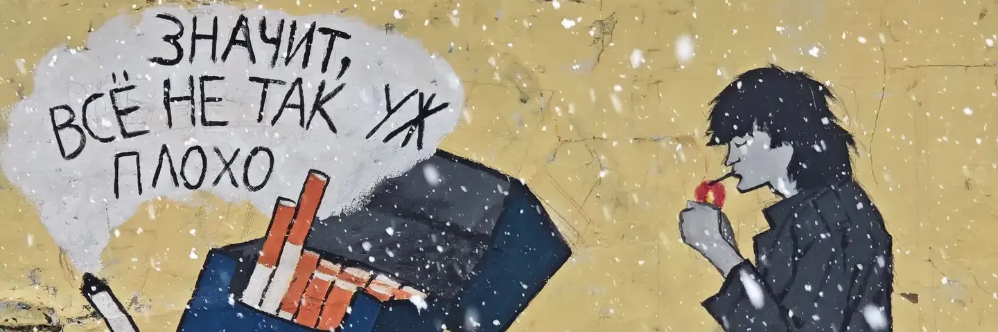 在一面粗糙的米黄色墙面上，张贴着一幅街头艺术作品，画面左侧是一个倾斜打开的蓝色香烟盒，其中一根香烟伸出盒外。烟盒上方有一团白色的云状烟雾，里面写着黑色的俄文句子“ЗНАЧИТ, ВСЁ НЕ ТАК УЖ ПЛОХО”。画面右侧是一个穿着深色衣服的黑色剪影人物，正点起一支烟，背景中有白色的雪花飘落。